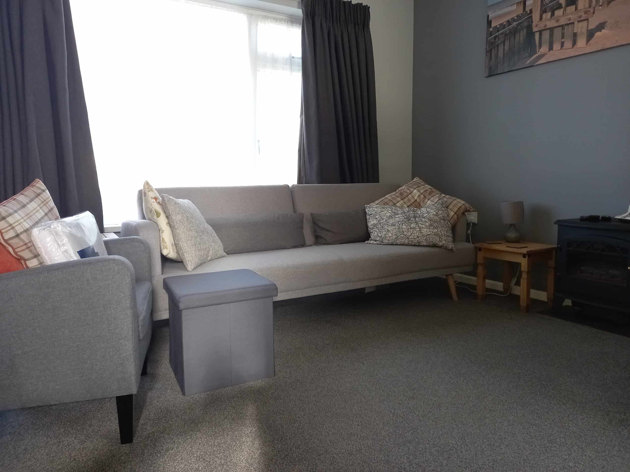 A cosy living room with a grey sofa, armchair, and ottoman. Cushions are arranged on the seating. There are dark curtains, a small side table with a lamp, and a wall art piece above the sofa. The carpet is grey.